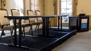 A short platform elevates two tables and four chairs into a stage-set-up. The stage is at the front of the room (near the projector screen) and next to the lectern.