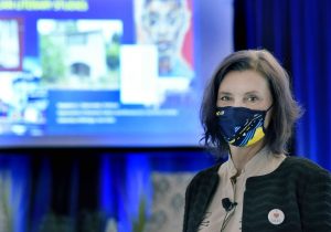 Magdalena Zaborowska, wearing a University of Michigan mask, stands in the University Room. Out of focus behind her, is her slideshow presentation.