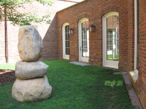 Part of The Conversation by Thomas Sayre: three large stones are stacked atop each other in the grassy courtyard outside of Hyde Hall, along the Fellows Gallery