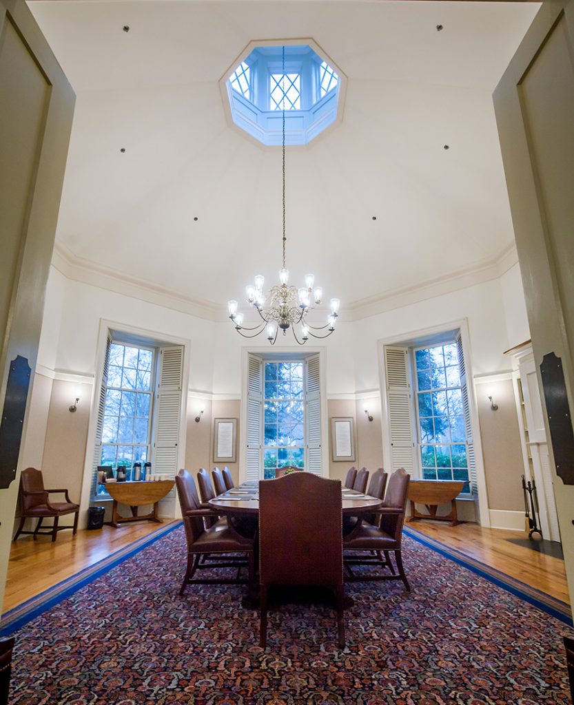 The doors propped open to show the Fellows Room, lit up from natural light and from the chandelier and sconces. Several tall-back leather chairs around the center table, and a red and blue-bordered rug is on the floor.