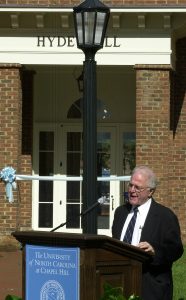 Ruel Tyson speaks at a lectern in front of the campus-facing entrance of Hyde Hall. Behind him, the entrance way is roped off with a Carolina blue ribbon.