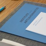 A blue folder and a white notepad with the Institute for the Arts and Humanities wordmark are set on table next to a pen and silverware.